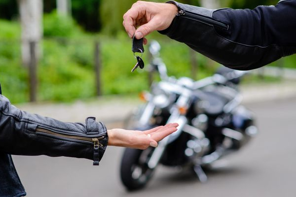 One person hands motorcycle keys to another, with a bike blurred in the background.